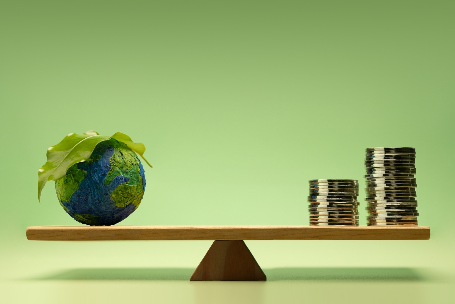 Altalena concettuale su sfondo verde con monete da un lato e il pianeta con una foglia dall’altro, simbolo dell’equilibrio tra finanza e sostenibilità